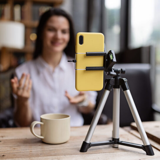 Happy young Caucasian millennial or gen z woman with long brunette hair streaming with smart phone on tripod, shooting social media blog in modern cafe. Influencer using social networks indoor. High quality photo