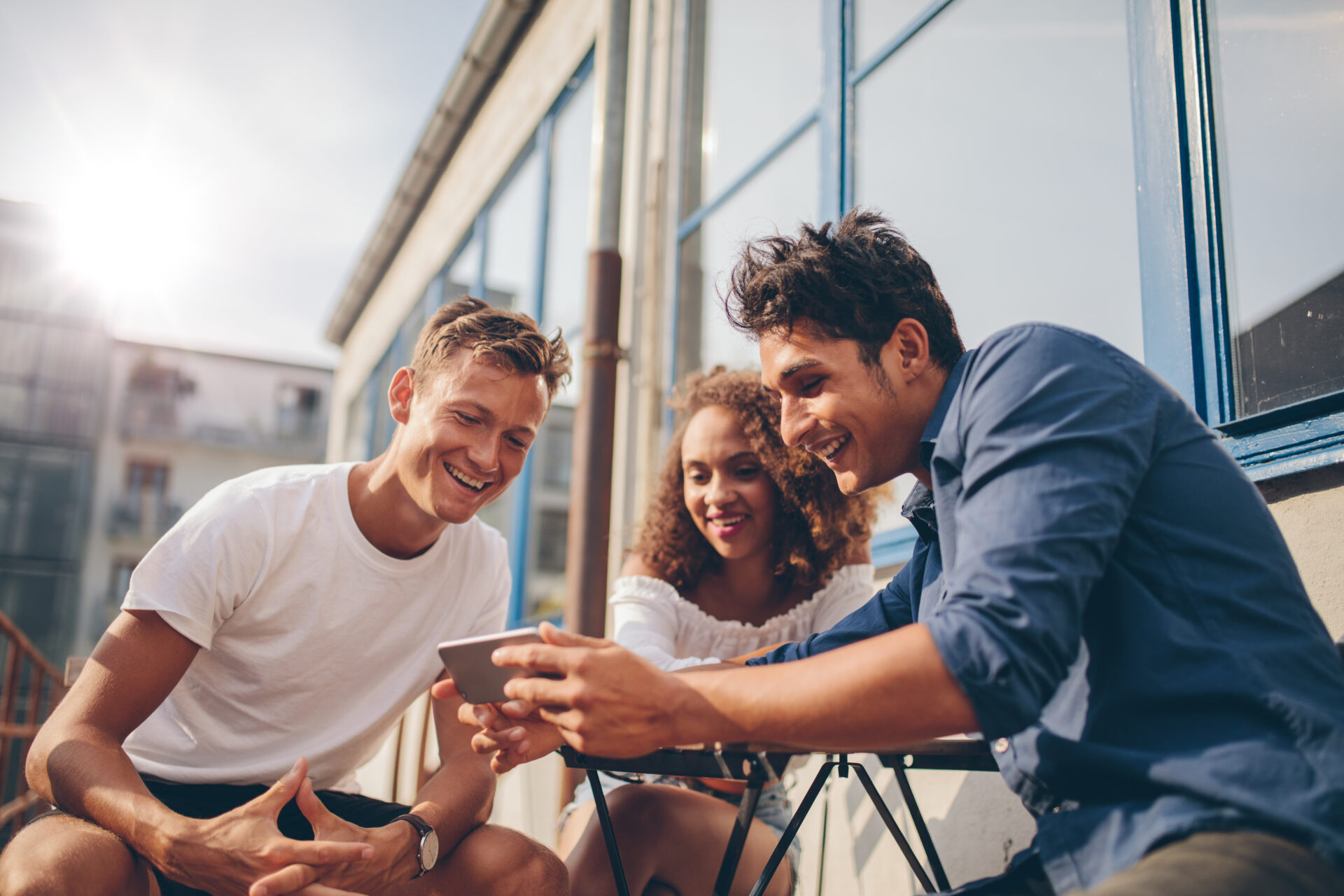 Three young friends sitting outdoors and looking at mobile phone. Group of people sitting at outdoor cafe and watching video on the smartphone.