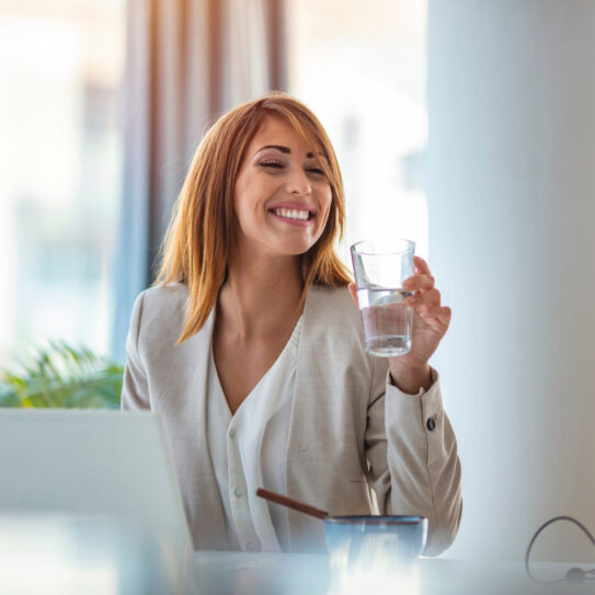 Businesswoman drinking water while working in her office. Close up of woman using a computer while holding a glass of water. Dehydrated female office worker drinking still mineral water