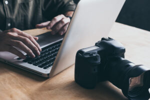 Close-up of a photographer on their laptop with their camera positioned on the desk behind the computer. 