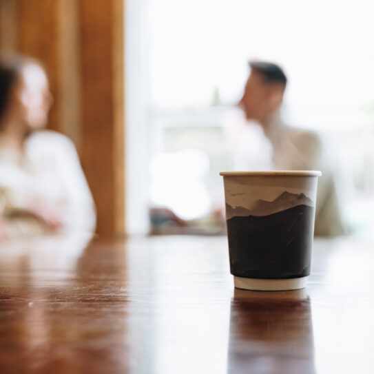 A close-up view of a coffee cup takes the center stage on a wooden table with a blurred background where two people appear to be engaging in a casual conversation in a cozy, indoor setting.